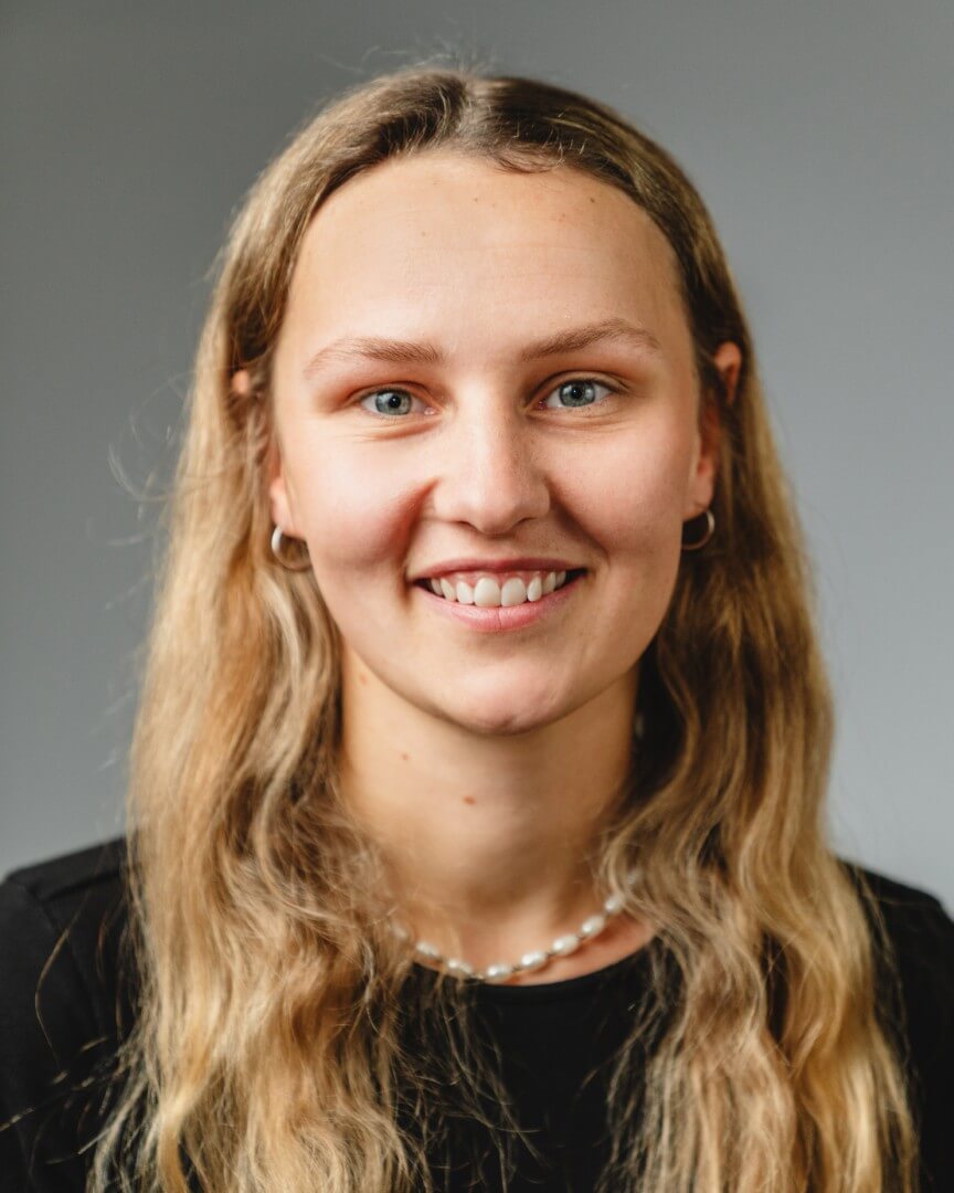 Person mit langen blonden Haaren lächelt vor grauem Hintergrund.