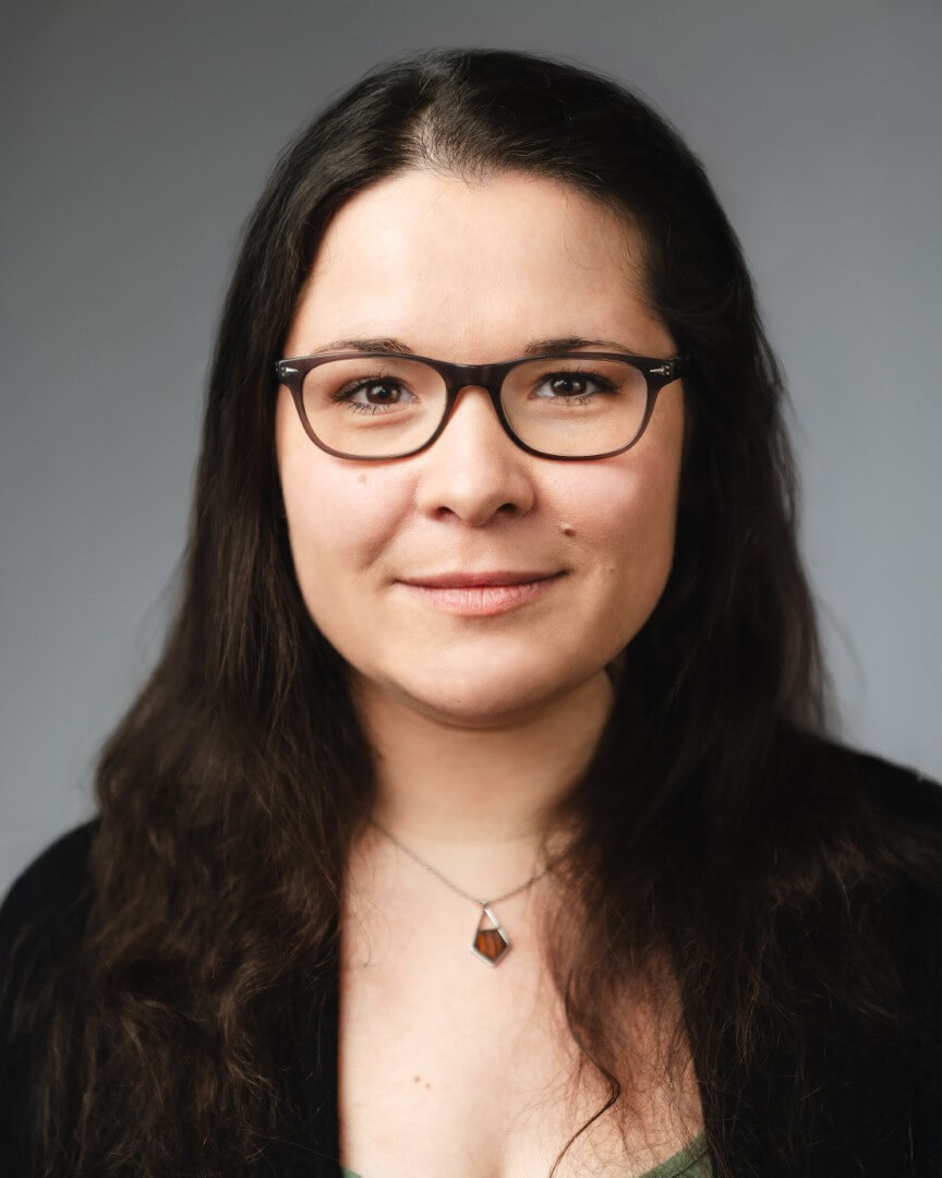 Frau mit Brille und langen Haaren trägt Kette, sitzt vor grauem Hintergrund.