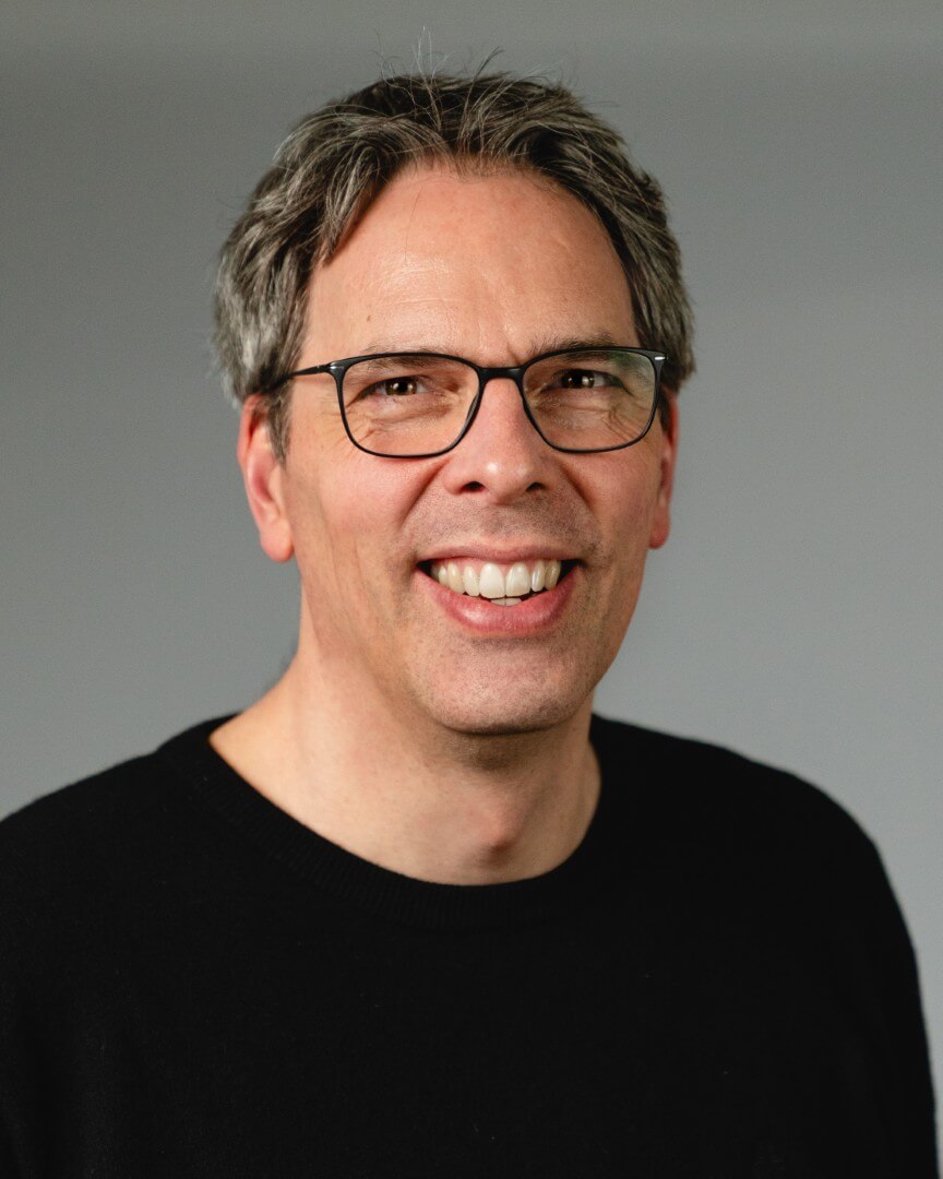 Lächelnde Person mit Brille und dunklem Pullover vor grauem Hintergrund.